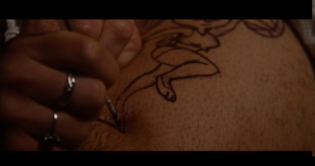 A close cropped shot showing a pair of hands decorated with rings firmly grasping a syringe and injecting deep into what appears to be a thigh with a tattoo of Lola Bunny playing basketball. The image highlights the realistic practical effect of a needle being plunged into someone's skin
